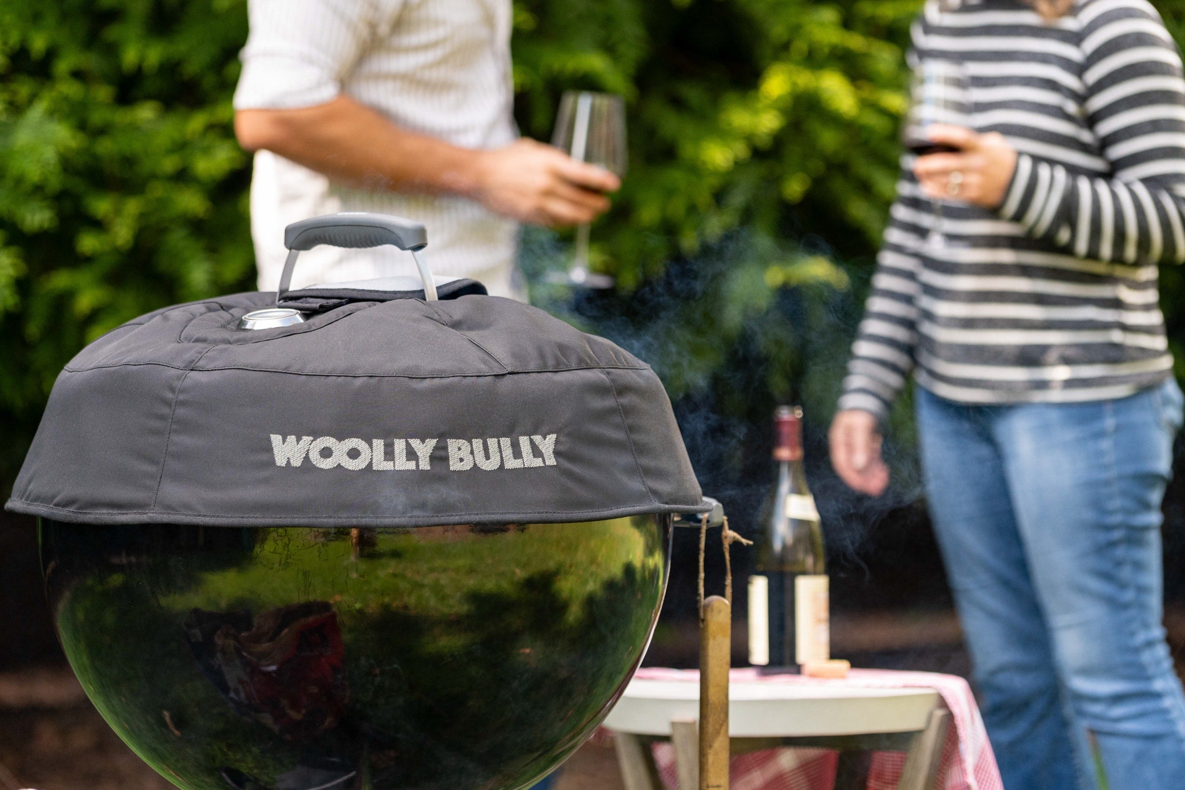 Two people relaxing in a backyard as they cook a meal on a Weber kettle with Woolly Bully Insulated Grill Cover